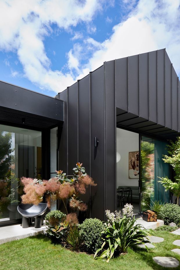 A contemporary dark-clad residential facade with pronounced vertical fins, projecting roof and entry canopy, and extensive glazing. A landscaped front garden with ornamental planting, stepping stones, and sculptural outdoor decor frames the entrance. The bright sky and strong black-and-green palette create a crisp, modern architectural presentation.