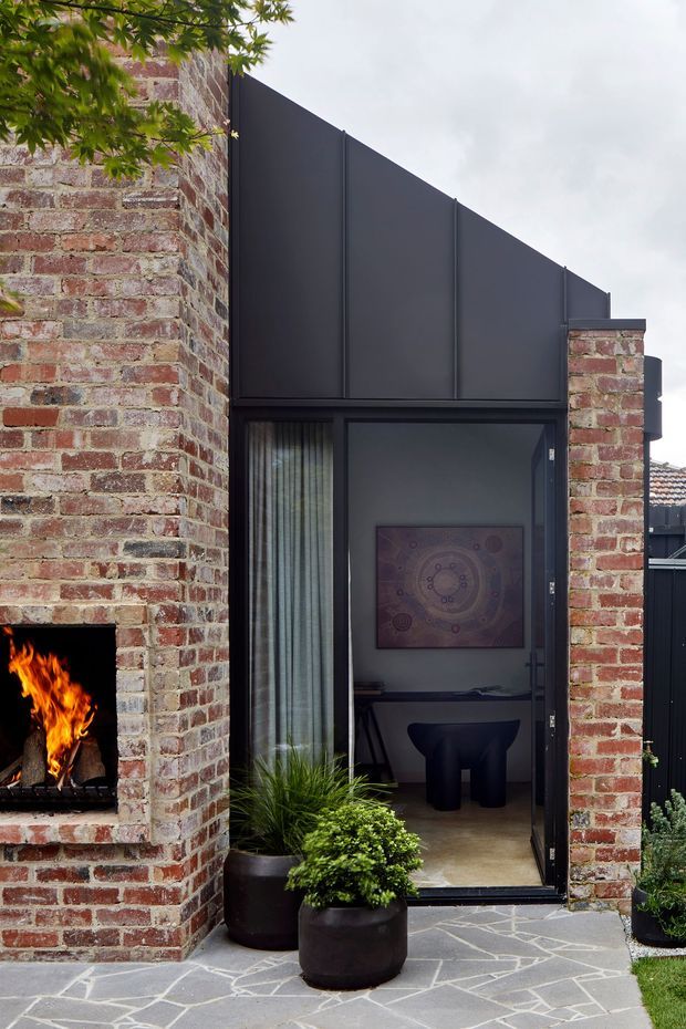 Exterior view of a contemporary brick extension or courtyard facade. A tall dark metal-clad gabled wall sits between exposed red-brick walls, with black-framed full-height glazing and a glazed door opening into a compact interior workspace or sitting area. The brick wall incorporates a built-in open fireplace with a visible fire. Irregular grey stone paving, potted plants, and mature foliage frame the entry.