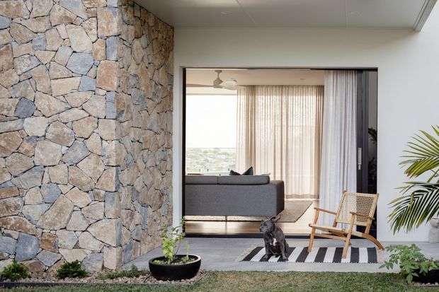 Contemporary residential interior/exterior threshold featuring a prominent irregular natural stone feature wall, smooth white ceiling and walls, and a wide opening into a bright living room. The living room contains a grey sofa, woven timber lounge chair, sheer curtains, a striped rug, and a ceiling fan, with indoor planting and a small dog visible. The composition also shows a landscaped foreground and a partial view toward a distant landscape through the glazing.