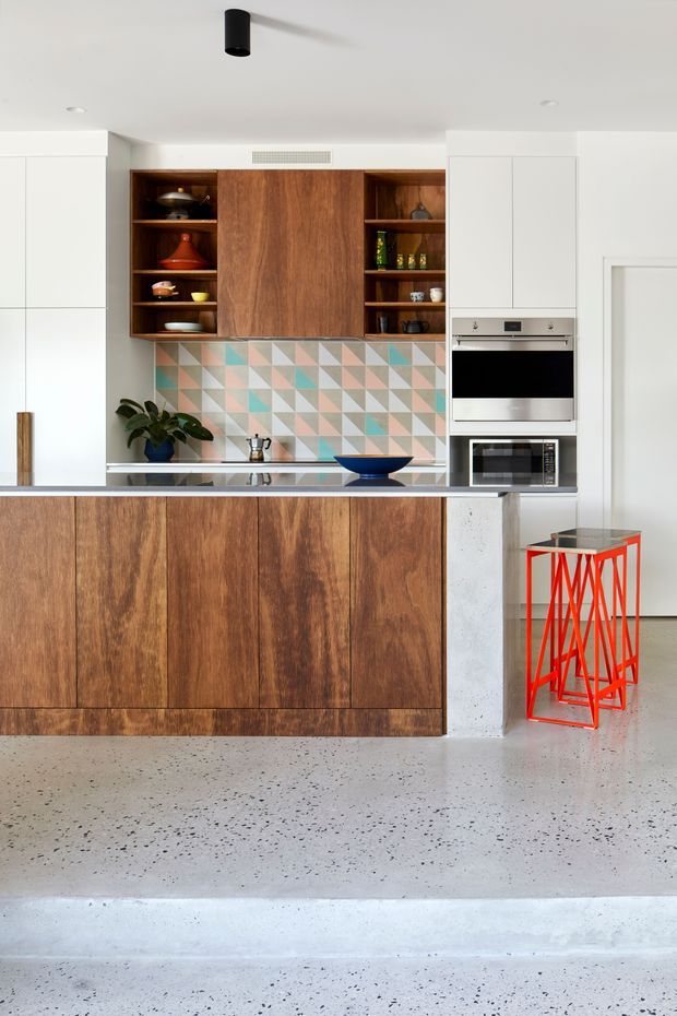 A bright contemporary kitchen with warm timber cabinetry, open timber shelving, white full-height cupboards, integrated stainless-steel oven and microwave, and a colourful geometric tiled splashback in pastel pink, mint, beige and grey. A dark benchtop, blue bowl, potted plant and vivid red geometric bar stool add accents. The kitchen opens onto a polished terrazzo or speckled concrete floor with a small level change.