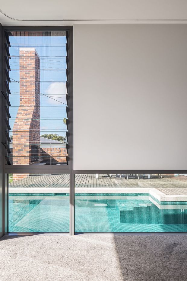 Interior view of a minimally finished room with a large floor-to-ceiling glazed opening overlooking a turquoise tiled swimming pool and timber deck. Dark aluminium window frames and external louvres create strong vertical and horizontal lines, while a brick wall and neighbouring roof are visible beyond. The room has pale carpet, smooth light walls, and abundant natural light.