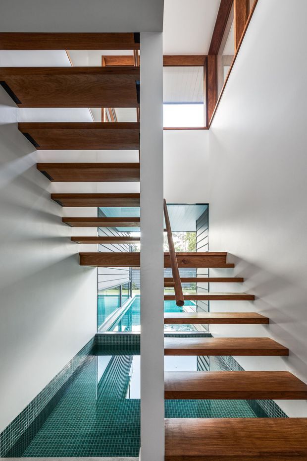 Interior architectural view of an open-riser timber staircase wrapping around a tall central white structural wall. Rich brown timber treads and handrail contrast with white walls and ceilings, while large windows and glazed openings bring daylight into the stairwell. A turquoise tiled pool or water feature is visible below and beside the stairs, creating a dramatic multi-level circulation space.