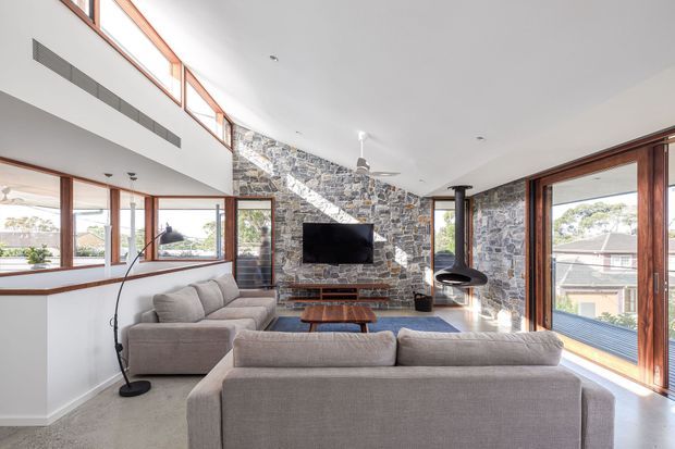 Bright, spacious contemporary living room with a double-height ceiling and extensive timber-framed glazing. A large sectional sofa arrangement faces a wall-mounted television set against a prominent natural stone feature wall, with a suspended black fireplace and timber coffee table adding focal points. The room opens visually toward exterior residential views and is filled with natural light.