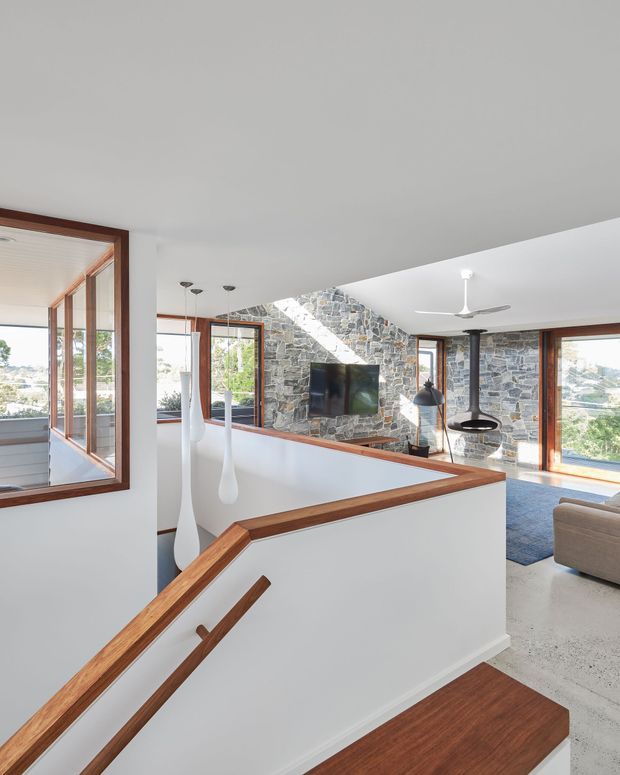 Bright contemporary split-level interior showing a stairway and landing with white walls, warm timber handrails and trim, polished light flooring, extensive timber-framed glazing, and a prominent exposed irregular stone feature wall. A living area with sofa, television, pendant lighting and ceiling fan is visible beyond the landing.