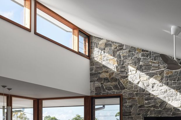 Interior architectural view featuring a tall stone feature wall, extensive timber-framed windows, and a partially visible double-height space with a mezzanine or upper-level opening. Natural daylight creates strong geometric shadows across the rough stone masonry. The palette is restrained and earthy, with white plaster, dark grey stone, and warm timber frames.