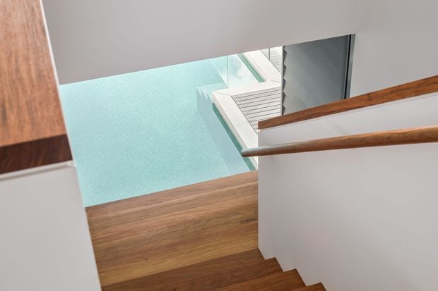 A contemporary stairway viewed from above, with dark timber stair treads and matching timber handrails set against crisp white walls. A large glazed opening beside the stairs provides a view toward a pale turquoise outdoor surface, creating a bright, geometric composition.