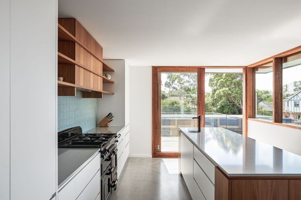 A bright contemporary kitchen with a long central island, white flat-panel cabinetry, warm timber open shelving and island joinery, pale blue-green tiled splashback, and a large stainless-steel benchtop. Full-height timber-framed glazing and a glazed door open to leafy suburban surroundings, providing strong natural light.
