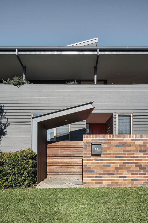 Front exterior view of a contemporary multi-level residence. The facade combines horizontal grey cladding, a projecting angular entry canopy, timber slatted gate, dark metal detailing, and a low red-brown brick boundary wall with a house number. A deep upper-level balcony or verandah is visible beneath a broad roof overhang, with a small roof element above. Neatly maintained lawn and shrubs frame the entrance.