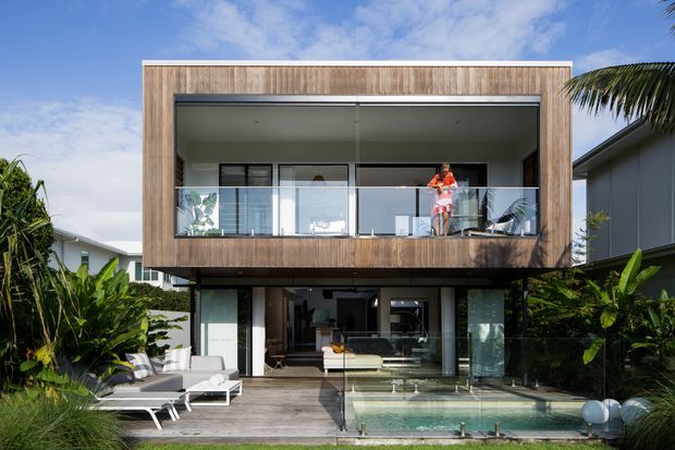 Contemporary two-storey residence viewed from the garden, featuring a projecting upper-level box clad in warm timber, extensive glazed openings, a glass-balustraded balcony, and a ground-level outdoor entertaining area with a small pool. Lush tropical planting surrounds the timber deck and pool.