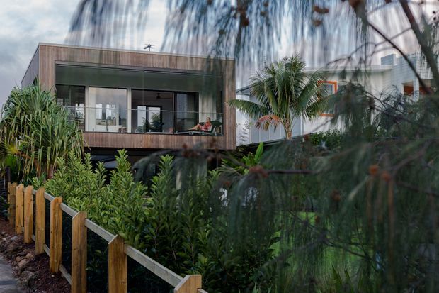 Exterior view of a contemporary multi-level residence partially screened by dense tropical and native planting. The building features a rectangular box form, expansive glazing, dark window frames, and warm timber-look cladding, with a balcony or terrace visible along the upper level. A timber-and-wire garden fence runs through the foreground.