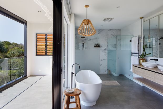 Bright contemporary bathroom with a freestanding white oval bathtub, floating timber vanity with a white vessel basin, black tapware, frameless glass shower enclosure, and large marble-look wall tiles. The room opens onto a glazed outdoor balcony or terrace, with a sculptural timber pendant light and mirrored cabinetry adding visual interest.