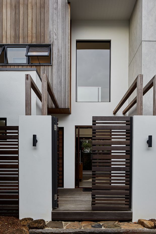 Front exterior view of a contemporary multi-level residence, focused on a recessed entry courtyard. The facade combines vertical weathered timber cladding, smooth white rendered walls, dark timber slatted privacy gates and fencing, slim dark-framed glazing, and a stone-paved threshold. An upper balcony and stair elements are partially visible, creating a layered, modern entrance composition.