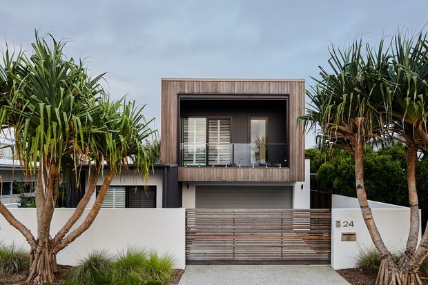 Front exterior view of a contemporary multi-level residence, featuring a box-like upper storey clad in vertical timber-look boards, dark recessed glazing, a ground-level garage, and a horizontal timber slat driveway gate. Tropical architectural planting frames the white rendered facade.