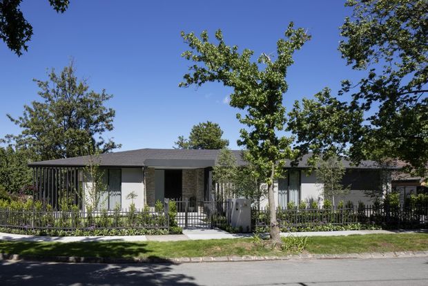 Street-facing view of a low, single-storey contemporary residence with a broad shallow-pitched dark roof, light rendered wall sections, dark vertical cladding or screening, extensive glazing, and a landscaped front garden behind a metal fence. Mature trees and a paved street frame the suburban setting.