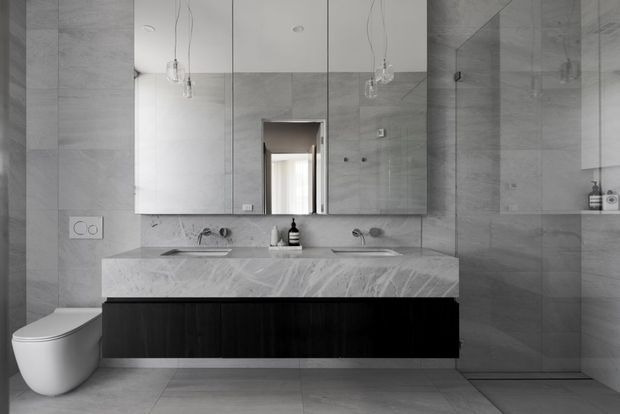 Contemporary monochromatic bathroom with large-format grey stone-look wall and floor tiles, a floating dark timber vanity with a broad veined stone benchtop, twin basins, mirrored cabinetry, wall-hung toilet, and a frameless glass shower enclosure. Minimal pendant lights and restrained fittings create a refined, clean appearance.
