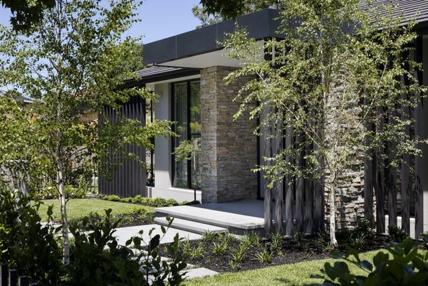 Contemporary single-storey home facade viewed from a landscaped front garden, featuring dark vertical cladding, extensive charcoal metal roof and trim, pale stacked-stone wall sections, large dark-framed glazing, and a covered entry with broad concrete steps. Established planting and lawn soften the modern exterior.