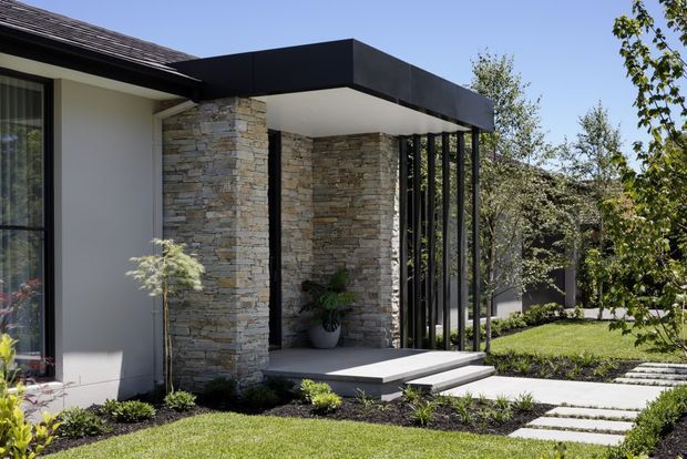 Contemporary residential entrance with a flat black canopy, stacked natural-stone feature walls, dark slender vertical screens, and a raised concrete entry platform. The facade is set within a landscaped garden with a stepping-stone path and lawn.