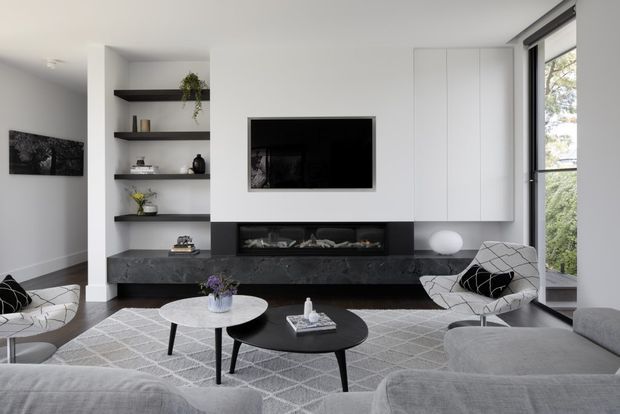 Contemporary living room with a wall-mounted television, built-in white cabinetry, open dark shelving, a long dark stone fireplace surround and hearth, and expansive corner glazing. The room is furnished with grey sofas, sculptural lounge chairs, nesting coffee tables, and a geometric grey rug.