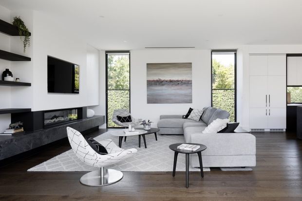 Bright contemporary living room with white walls, dark timber flooring, a large pale sectional sofa, sculptural lounge chairs, black nesting coffee tables, and a geometric light rug. Built-in black shelving and media cabinetry line the left wall, while tall windows provide abundant natural light and views of greenery. White built-in storage and a glimpse of the kitchen are visible at right.