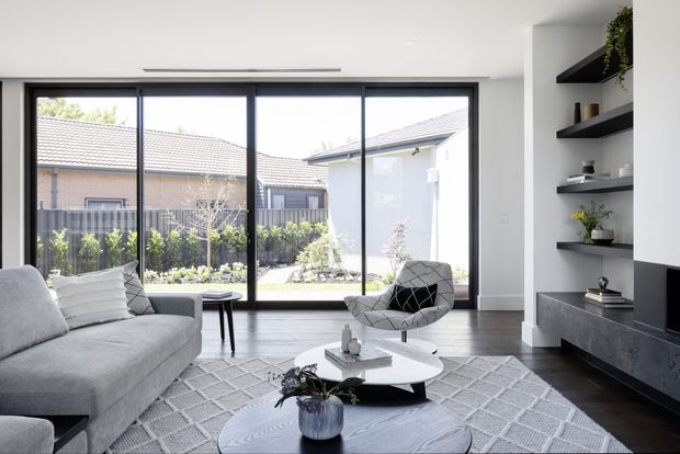 Contemporary living room with a large grey sectional sofa, sculptural lounge chair, layered round coffee tables, and a geometric area rug. Full-height black-framed glazing brings in abundant daylight and overlooks a landscaped courtyard and neighbouring homes. Built-in dark shelving and low media cabinetry line the right wall, with a clean white ceiling and dark timber-look flooring.