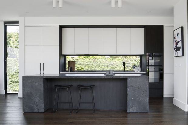 A refined contemporary kitchen with a large central island, dark stone sides and benchtop, dark timber-look cabinetry, and two black bar stools. White full-height cabinetry surrounds integrated appliances, while a long horizontal window above the sink brings in greenery and natural light. The room has dark timber flooring, crisp white walls, black-framed glazing, and minimalist black-and-white artwork.