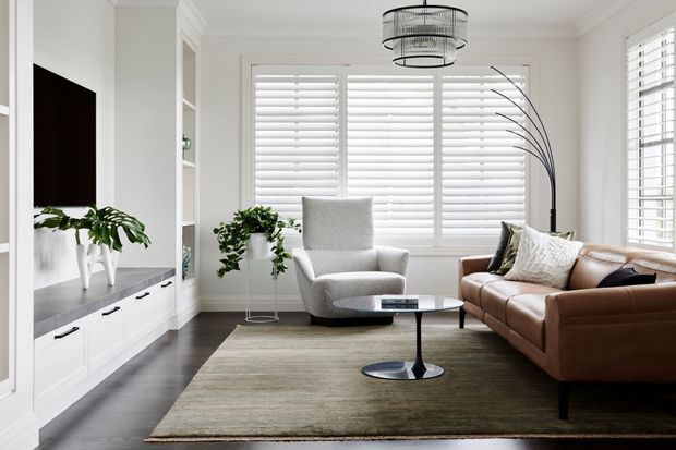 Bright, refined living room with white built-in media cabinetry, a wall-mounted television, a grey upholstered lounge chair, a brown leather sofa, a round coffee table, indoor plants, a large area rug, and extensive white plantation shutters. A contemporary circular pendant light and sculptural floor lamp add visual interest.