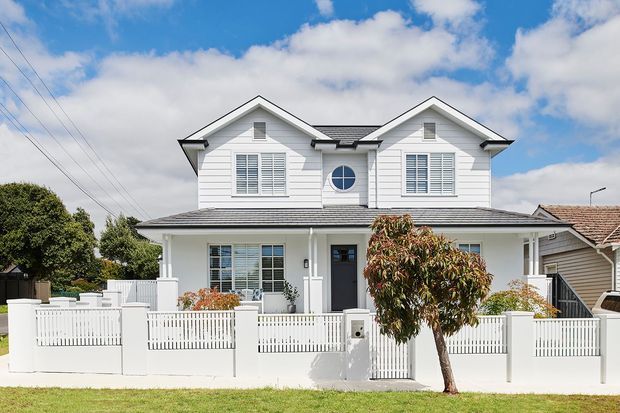 Street-facing exterior view of a two-storey white weatherboard home with a symmetrical twin-gable facade, dark tiled roof, shuttered windows, central circular window, covered front porch, and white picket-style boundary fencing. A neighboring house and overhead utility lines are visible in the background.