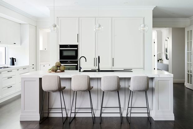 Bright, spacious white kitchen with extensive shaker-style cabinetry, a large central island with seating for four, integrated double ovens, pendant lights, dark timber flooring, and a black gooseneck tap. The room has a refined, clean and contemporary-classic appearance with strong white and light-grey tones contrasted by dark flooring and fixtures.
