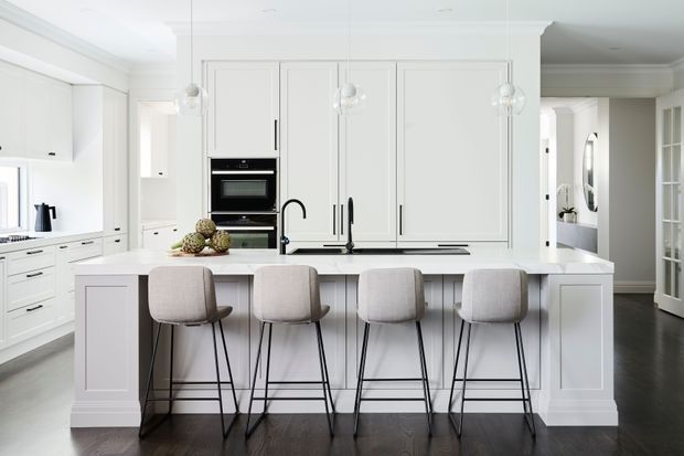 Bright, refined white kitchen with a large central island, four upholstered bar stools, integrated cabinetry, built-in black ovens, dark timber flooring, and pendant globe lights. The island includes a sink and black tap, while a glass-paneled door and adjacent hallway are visible in the background.