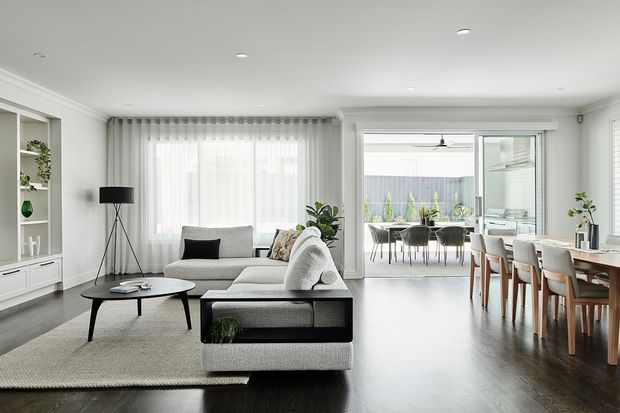 Bright open-plan contemporary living and dining area with a neutral palette, dark timber flooring, light upholstered sectional sofa, round black coffee table, built-in white shelving, and a large dining table. Full-height glazed doors open to an outdoor dining terrace or courtyard, with abundant natural light and sheer curtains.
