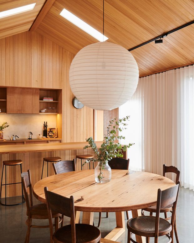 Warm, wood-lined open-plan dining and kitchen area with a round solid timber dining table surrounded by vintage-style wooden chairs. A timber kitchen and breakfast bar with stools sits behind the dining area, while a large paper globe pendant, linear skylights, track lighting, indoor greenery, and full-height sheer curtains add softness and natural light.