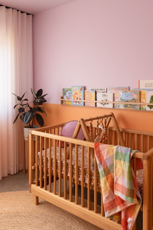 A warmly styled nursery with a natural timber cot in the foreground, patterned bedding, a wooden baby mobile, and a colourful checked blanket draped over the rail. The room has a two-tone pink and terracotta wall treatment, pale curtains, a woven rug, a rubber plant, and a narrow wall-mounted display shelf filled with children's books. Soft daylight creates a cosy, playful atmosphere.