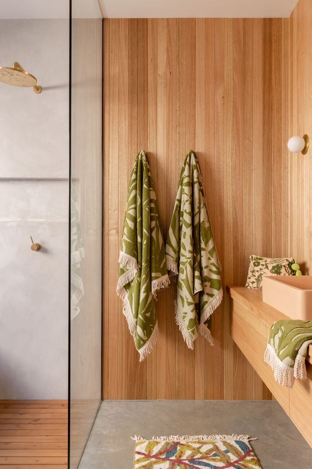 Warm contemporary bathroom with a walk-in glass shower on the left and a timber-lined vanity wall on the right. Vertical light timber wall panelling, a concrete-look floor, brass/gold fittings, and patterned green textiles create a natural, relaxed feel.