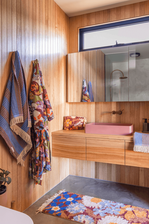 A compact, richly styled bathroom featuring vertical timber wall panelling, a floating timber vanity with integrated drawers, a pink vessel basin, wall-mounted bronze-toned tapware, and a large mirrored cabinet. A glass shower enclosure is visible in the reflection. Colourful patterned textiles and a vibrant geometric bath mat add a playful decorative accent.