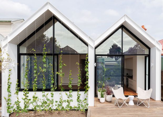 Exterior view of a contemporary pair of steeply gabled, white-clad glazed pavilions. Black-framed floor-to-ceiling glazing opens onto a timber deck, with climbing plants along the facade and a glimpse of a minimally furnished interior.