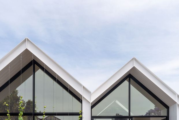 Exterior architectural view of a contemporary building with two prominent steep gabled roof forms, white trim, dark metal framing, and large floor-to-roof glazing. The image focuses on the facade and roofline against a pale sky, with minimal landscaping visible at the base.