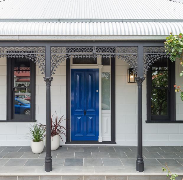 Front elevation of a traditional single-storey home featuring a covered verandah, ornate dark metal lacework and slender columns, white weatherboard-style cladding, a vivid blue paneled entry door, dark-framed windows, and grey tiled paving. The corrugated metal roof and decorative porch detailing create a heritage character.