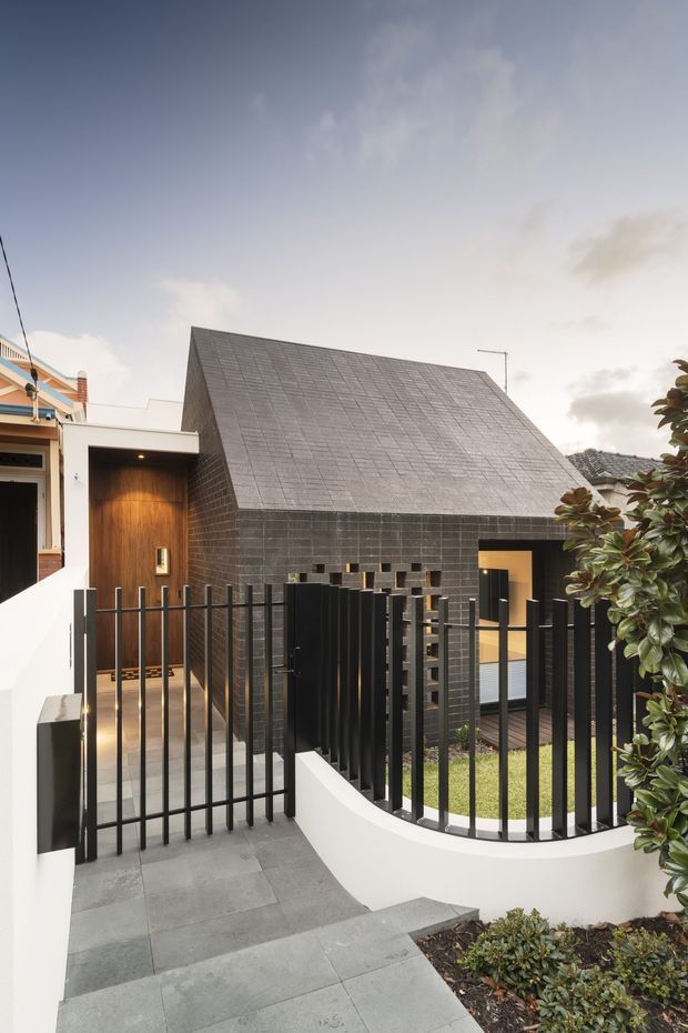 Exterior architectural photograph of a compact contemporary house viewed from the front walkway. The home features a steep gabled roof and facade clad in dark grey rectangular tiles, a warm timber entry wall, black metal fencing and gates, and a curved white rendered boundary wall with a landscaped garden. A neighbouring dwelling is partially visible at the left.