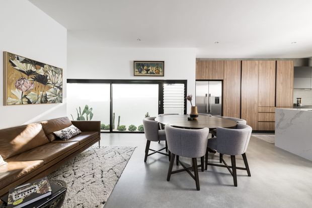 Open-plan contemporary living, dining and kitchen space with white walls, polished concrete flooring, extensive glazing, a brown leather sofa, round dining table with upholstered chairs, and warm timber kitchen cabinetry. The room is bright and clean with a restrained neutral palette and natural light.