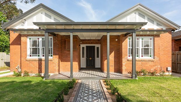 Front exterior view of a single-storey brick home with two prominent gabled roof forms, white rendered gable ends, white plantation-shuttered windows, and a covered entry porch. A patterned tiled path leads through the front lawn to the central entrance, with timber fencing and garden planting at the sides.