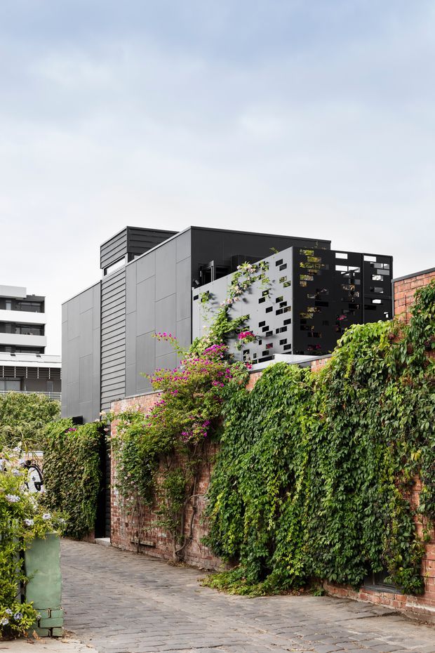 Exterior view of a contemporary multi-storey urban building behind an old brick wall covered in dense climbing greenery. The building has a sharply rectilinear form, dark and light grey panelled façades, horizontal louvred cladding, and perforated screen-like sections with planted openings. A paved pedestrian lane runs alongside the landscaped brick boundary.