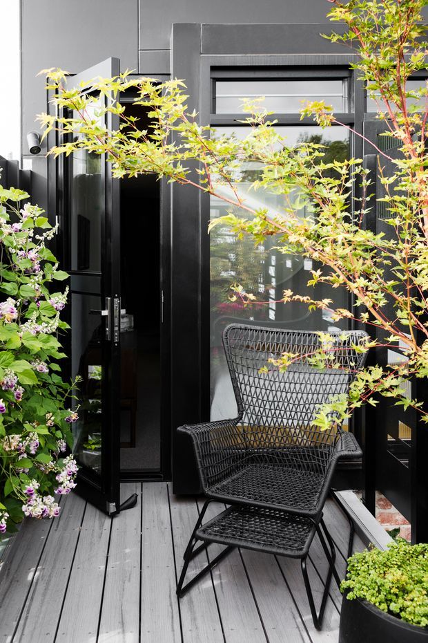A compact contemporary outdoor terrace or balcony with dark charcoal cladding, black-framed glazing and an open glass door. Grey timber decking supports a black woven lounge chair and footstool, while leafy potted plants and flowering greenery soften the monochromatic facade.