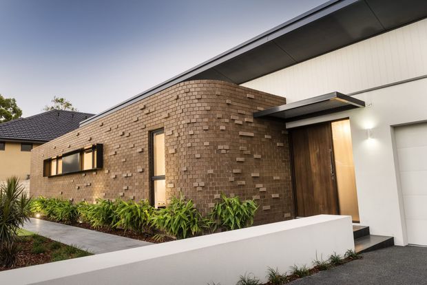 A contemporary single-storey house exterior at dusk, featuring a dark textured brick facade with projecting brick detailing, white rendered walls, a low-pitched dark roof, timber front door, narrow vertical glazing, and a white boundary wall. Landscaped planting lines the concrete path and entrance.