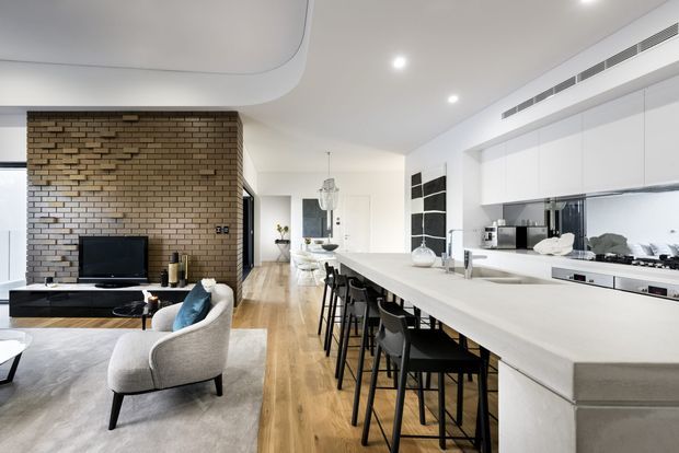 Open-plan contemporary apartment interior combining a sleek white kitchen with a long island and bar seating, a dining area, and a living room. The space features warm timber flooring, a textured exposed-brick feature wall, white streamlined cabinetry, recessed lighting, and a sculptural curved ceiling detail.