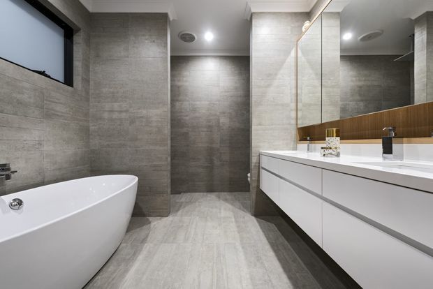 Contemporary bathroom featuring large-format grey stone-look tiles across the walls and floor, a freestanding white bathtub, frameless glass shower enclosure, and a long floating white vanity with integrated basins. A warm timber-toned backsplash panel and large mirrored wall add contrast and reflect the modern fittings.