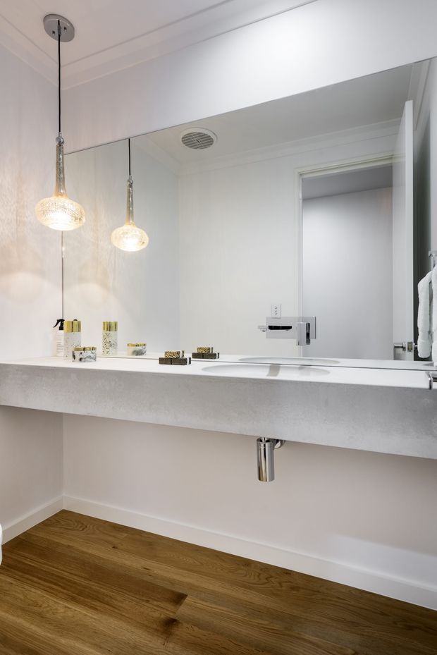 Bright, contemporary bathroom vanity viewed straight on, featuring a long pale stone or engineered-stone benchtop with an integrated basin, wall-mounted chrome tapware, a large mirror, white walls and trim, pendant lights, and warm timber flooring. A towel and open doorway are visible at the right.