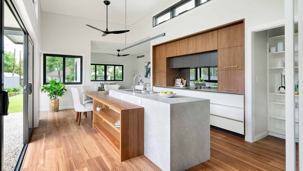 Bright open-plan kitchen and dining area with a large waterfall-edge island, integrated white cabinetry, and a warm timber feature wall framing the cooktop and rangehood. The room has timber flooring, high ceilings with black ceiling fans, clerestory windows, extensive black-framed glazing, and an adjacent built-in open pantry or shelving area. A dining table and living area are visible beyond the kitchen.