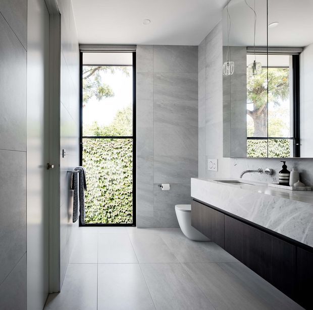 A bright, contemporary bathroom with large-format grey stone-look wall and floor tiles, a floating dark timber vanity with a pale stone benchtop, wall-mounted tapware, a white toilet, frameless mirror, black-framed window, and minimalist pendant lights. Natural greenery is visible outside the window.