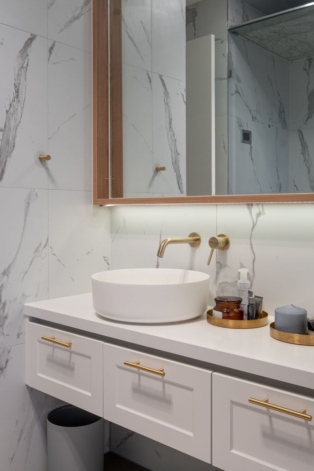 Close-up view of a refined bathroom vanity with a white vessel basin, wall-mounted brushed gold tapware, white shaker-style cabinetry with gold handles, and a large timber-framed mirrored cabinet. The walls are clad in white marble-look tiles with prominent grey veining, complemented by under-vanity lighting and gold-toned bathroom accessories.