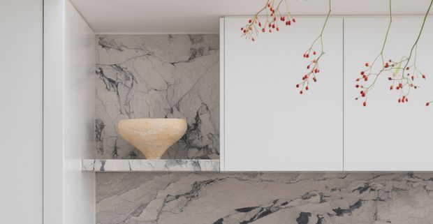 Close-up of a refined minimalist bathroom vanity or display joinery. White flat-panel cabinetry and a continuous veined grey-white marble surface frame a recessed niche containing a sculptural beige vessel basin. Delicate branch-like decorative elements hang in the foreground, adding a restrained organic accent.